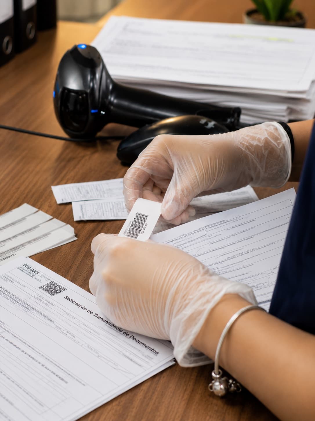Mãos com luvas aplicando uma etiqueta de código de barras sobre uma Solicitação de Transferência de Documentos da SOS Docs, com o leitor óptico e outras etiquetas na bancada