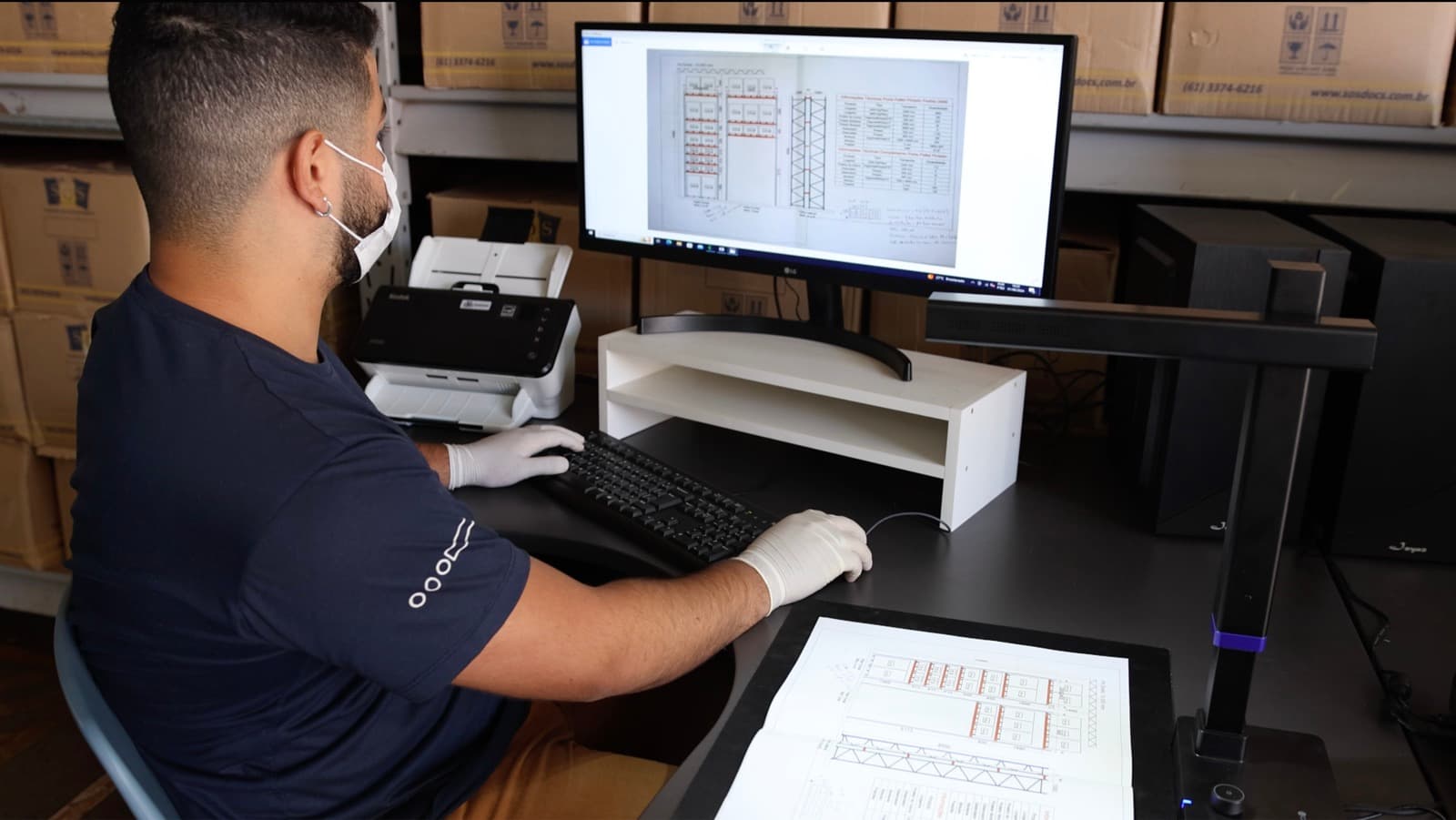 Operador da SOS Docs, de luvas e máscara, digitalizando uma planta técnica em estação de captura com scanner de mesa e scanner planetário, cercado por caixas de arquivo identificadas