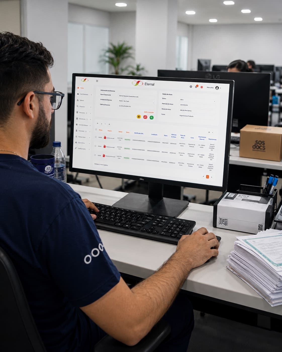 Profissional consulta o sistema Eternal em um monitor, ao lado de uma pilha de documentos e de uma caixa-arquivo da SOS Docs.